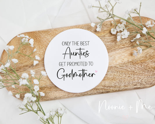 Round white ceramic coaster with text 'Only the best Aunties get promoted to Godmother' on a wooden board with flowers.