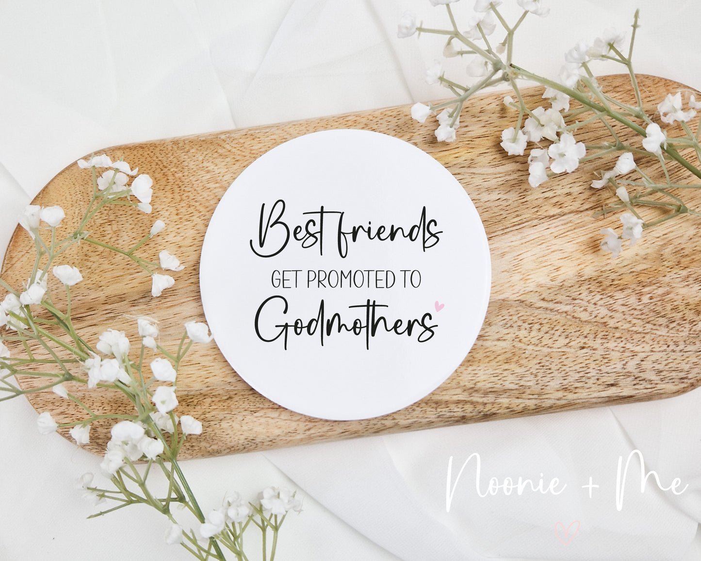Round white ceramic coaster with 'Best friends get promoted to Godmothers' text on a wooden board with white flowers.