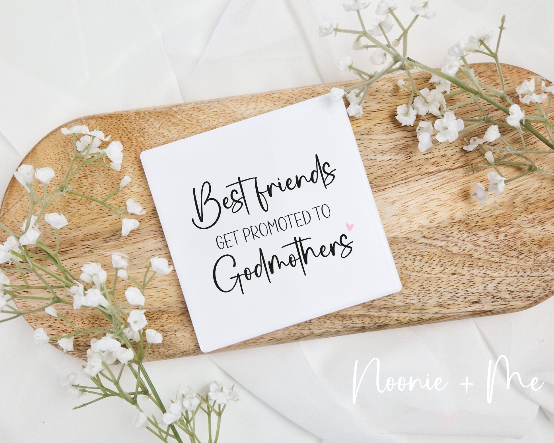Square ceramic coaster with 'Best friends get promoted to godmothers' text on a wooden board with flowers.