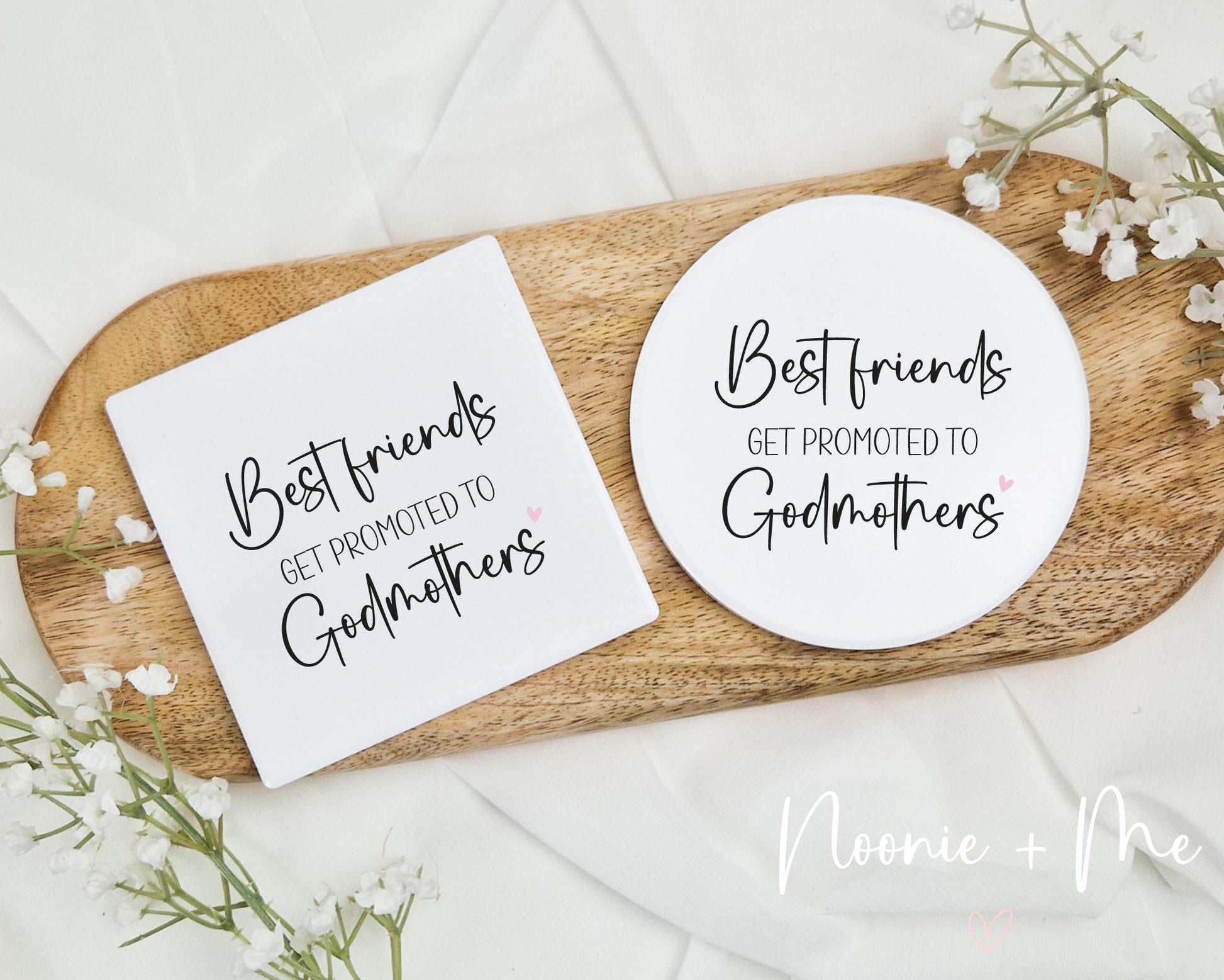 Two coasters with 'Best friends get promoted to Godmothers' text on a wooden tray with white flowers.