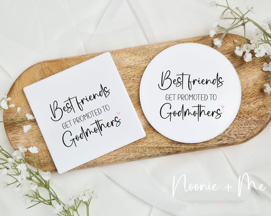 Two coasters with 'Best friends get promoted to Godmothers' text on a wooden tray with white flowers.