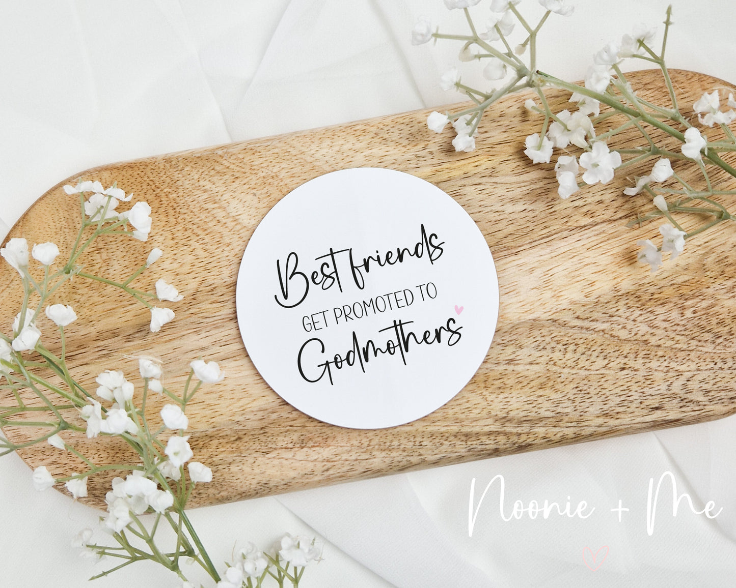 Round wooden coaster with 'Best friends get promoted to Godmothers' text on a wooden board with white flowers.