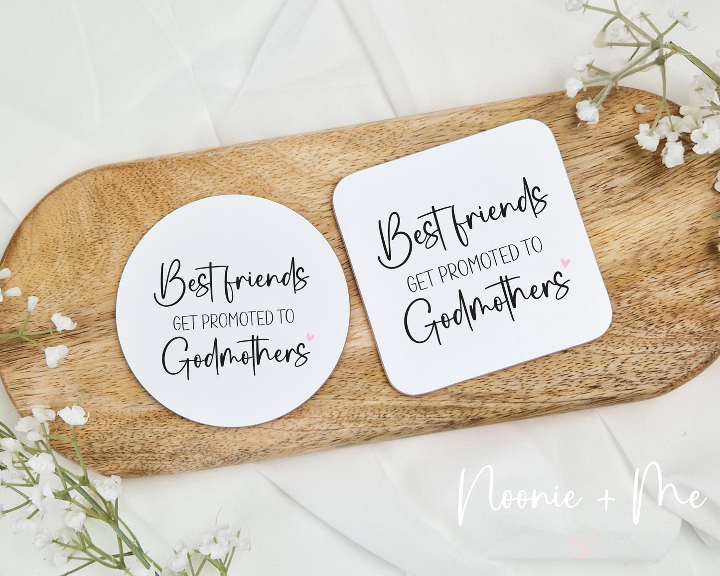 Two coasters with 'Best friends get promoted to godmothers' text on a wooden board with flowers.