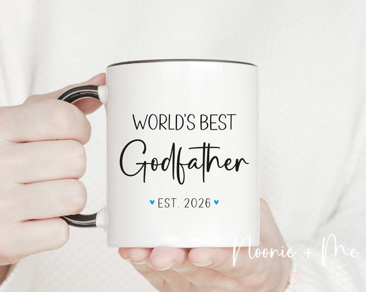 White mug with 'World's Best Godfather Est. 2026' text held by a hand against a white background.
